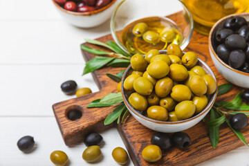 Green and black olives on a white wooden background. Various types of olives in bowls and olive oil with fresh olive leaves. Copy space. Place for text. Mediterranean food. Vegan.