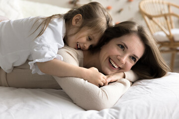 Fototapeta na wymiar Cheerful cute toddler girl resting on happy moms back, laughing, having fun. Happy mother enjoying motherhood leisure, lying on bed at home, piggybacking little kid, playing active game with daughter