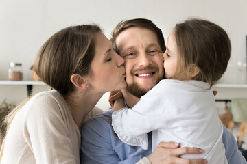 Pretty loving young mom and little daughter kissing happy dad celebrating fathers day, looking at camera, smiling, holding toddler child in arms, hugging wife, enjoying family leisure, shooting