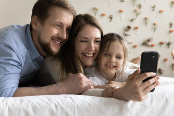 Happy young parents hugging little toddler kid, talking selfie on cellphone, posing for self picture together, speaking on online family video call, enjoying domestic Internet communication