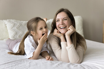 Cheerful caring mom lying at kid on double bed before yawning toddler girl falling asleep, talking with child, laughing, relaxing on spacious comfortable mattress with white sheet