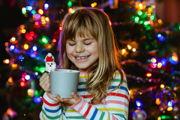 Little child girl holding cup with hot chocolate with marshmallows as Santa Claus. Kid sitting near Christmas tree decorated with lights. - Powered by Adobe