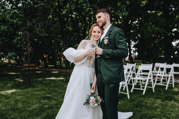 A stylish bearded groom and a beautiful smiling bride in a white dress are hugging, holding glasses...