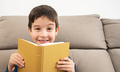 Boy sitting on sofa and holding book in front of his face at living room with copy space