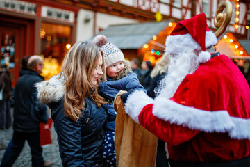 Little toddler girl with mother on German Christmas market. Happy kid taking gift from bag of Santa Claus. Smiling woman and daughter, family celebrating traditional holiday