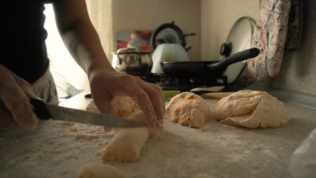 a woman in the kitchen rolls out a roll of dough and cuts it into small pieces with a long sharp knife. all around is in a mess. crockery in the background