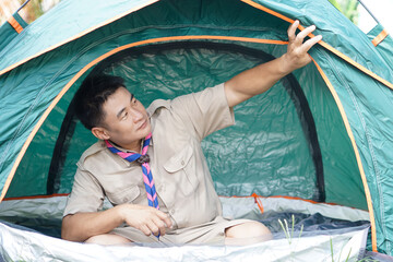 Handsome Asian man wears boy scout uniform sits in camping tent. Concept, accommodation for temporary outdoor sleeping as scout hiking activity. Scout go camping.         - obrazy, fototapety, plakaty
