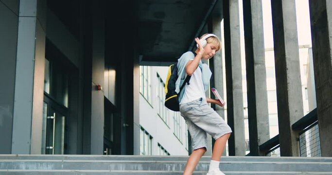 Handsome happy caucasian schoolchild with school bag and headphones listen to music walking down the stairs after school classes, outdoors