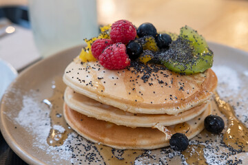Stack of thick, fluffy American-style pancakes served with maple syrup poured over fresh berries and kiwi, sprinkled with poppy seeds, delicious dessert plate easy to prepare for breakfast or brunch
