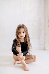 a cute little girl in a black gymnastic swimsuit is sitting on the floor in a white light hall, the...