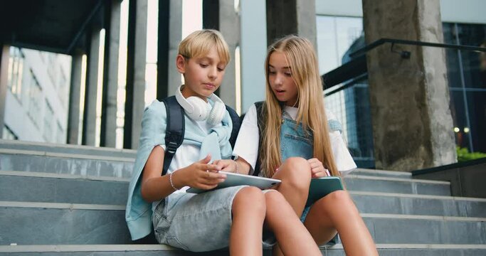 Positive teenage boy and a girl walking sittingding near school building on the stairs, having fun, talking about ppast lecture, use tablet deice at bright summer day, outdoors