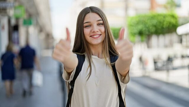 Young beautiful girl student wearing backpack doing thumbs up gesture at street