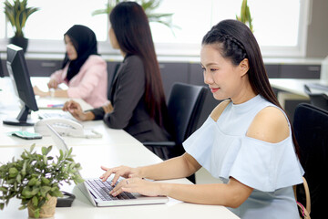 Portrait of happy beautiful Asian woman officer sitting at office desk, typing on laptop computer with blurred background of busy working colleagues. Pretty smiling female staff worker at workplace.