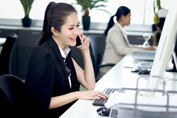 Happy smiling Asian woman officer sitting, talking on mobile phone at office desk with blurred busy colleague background, modern fashionable female businesswoman beautiful working at workplace.