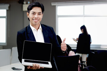 Portrait of happy smiling Asian businessman in black suit holds laptop computer and gives thumb up at office building with blurred background of colleagues, confident male officer working at workplace