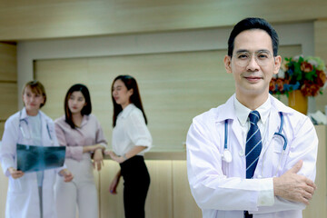 Portrait of smiling senior Asian male doctor in workwear with stethoscope, standing with arms crossed in clinic hospital, looks at camera and medical team working with patient as blurred background.