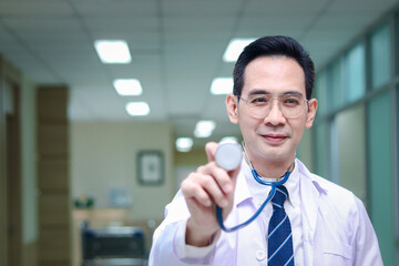 Portrait of friendly smiling senior Asian male doctor in white workwear showing stethoscope to camera, standing in corridor of medical clinic, happy doctor at hospital, medical and health care concept