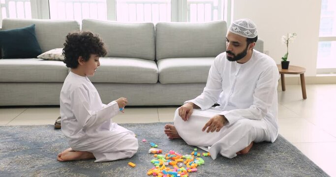 Muslim family spend time together play plastic blocks sit on floor in living room. Loving father in traditional Islamic clothes gives high five praising son enjoy playtime at home. Fatherhood, culture