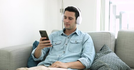 Hispanic man in wireless headphones holds smartphone choose podcast for listening use audio digital streaming media services provider resting on sofa spend leisure at home. Wireless, modern tech usage