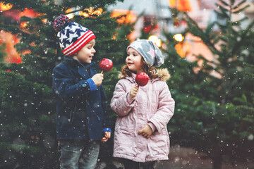 Two little smiling kids, boy and girl eating crystalized sugared apple on German Christmas market....