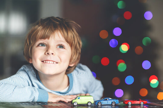 Little blond child playing with cars and toys at home, indoor. Cute happy funny kid boy having fun with gifts. Colorful lights on background. Christmas time concept..