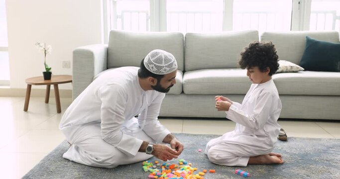 Muslim family spend time at home play plastic toy set seated on floor in modern living room, loving dad in traditional Islamic clothes enjoy playtime with preschooler little son. Fatherhood, culture
