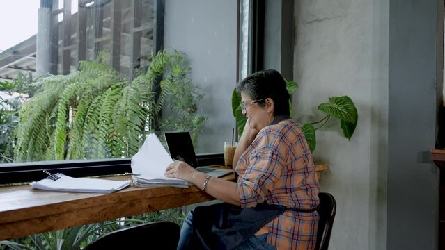 Elderly Asian mother is mother owner cafe bakery where sits, have happy smiling expression, when checking out shop account for daughter, get decent amount profit back shop, laptop computer on table.