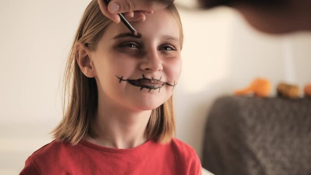 Mother making Halloween makeup to preteen girl daughter and painting spooky black lines on face. Pretty child with creepy cosmetics at October holiday death day celebration