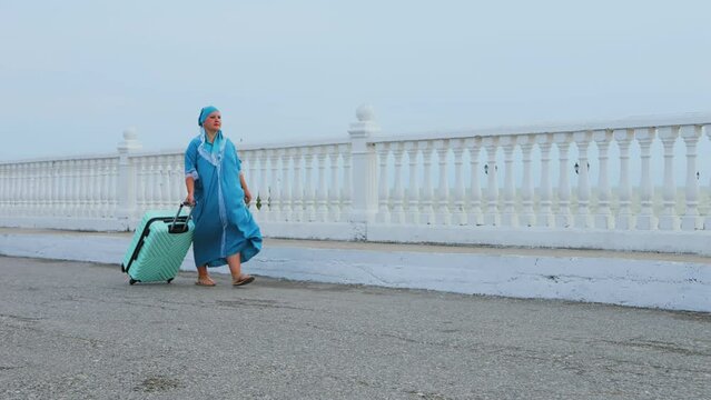 A Jewish woman in a turquoise dress and headscarf walks early in the morning with a suitcase along the embankment against the backdrop of the sea. Overall plan