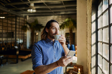 Young Caucasian man drinking coffee and using a smartphone in a startup company office
