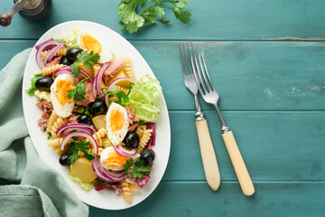 Tuna salad with pasta, eggs, potatoes, olives, red onions and sauce in white plate on old turquoise wooden rustic table background. Nicoise salad. French cuisine. Top view. Flat lay.
