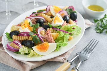 Tuna salad with pasta, eggs, potatoes, olives, red onions and sauce in white plate on old light gray concrete table background. Nicoise salad. French cuisine. Top view. Flat lay.