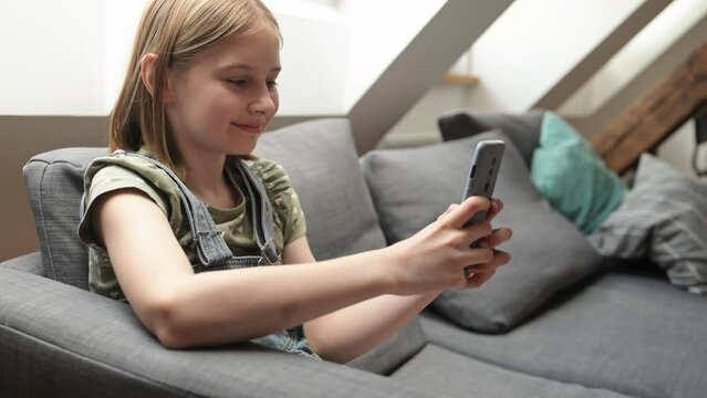 Child girl with smartphone sitting on sofa at home, chatting with friend in social media and smiling. Pretty preteen kid with mobile phone texting