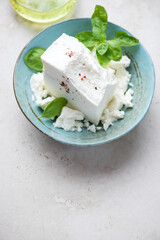 Turquoise bowl with a block of feta cheese and green basil on a light-beige stone background, vertical shot with space, selective focus