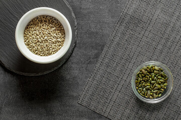 Sunflower and pumpkin seeds peeled in bowls on slate stone plate and grey tablecloth, dark background, top view