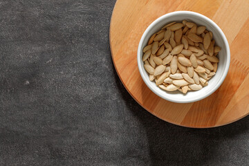 Pumpkin seeds, unpeeled with salt in bowl on wooden round board, on dark background, top view, space to copy text