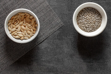 Pumpkin seeds unpeeled and sunflower seeds peeled in bowls, on dark background, top view, space to copy text