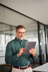 Smiling busy middle aged business man investor using tablet computer, mature older businessman executive manager looking at tab satisfied with finance trading market standing in office. Vertical