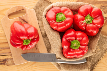Four sweet red peppers in a jute bag with a knife and a wooden kitchen board on a wooden table, macro, top view.