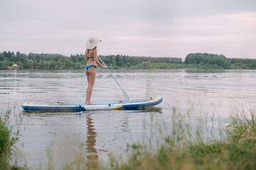 Fototapeta na wymiar A young woman with an open swimsuit swims on a SUP board on a picturesque lake. Substitute the paddle. A beautiful, slender girl is engaged in sap surfing on the calm water of a picturesque pond.