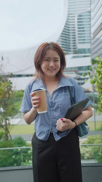 Cheerful young asian woman holding tablet and a cup of coffee smiling to camera at outdoors, city view. Young woman standing and looking at camera. Vertical