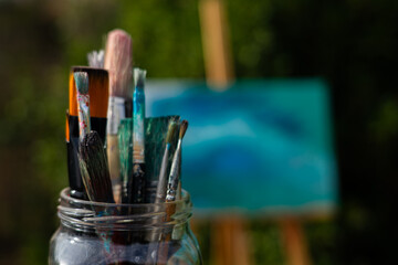 Many brushes in a glass jar with an easel with a painted canvas behind