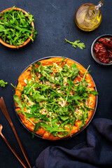 Hot pizza with mozzarella cheese, ham, pesto and fresh arugula, black table background, top view