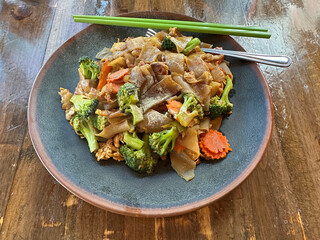 A pad see ew dish is shown at a Thai restaurant consisting of stir fried noodles, chicken, broccoli, carrots, and fried eggs.