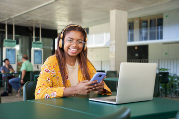 Portrait of a young black girl using headphones a phone and laptop. She looks at the camera and...