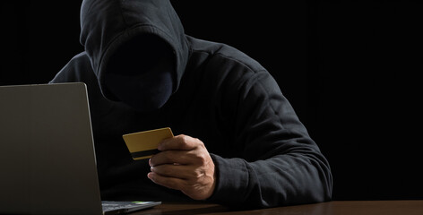 Hacker spy man one person black hoodie sitting on table hand holding credit card look computer...