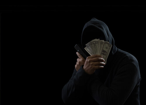 Portrait man thief wearing a black hood shirt, standing hand holding gun and money cash, counting the amount obtained from hijacking or robbing, in pitch-black room. dark background