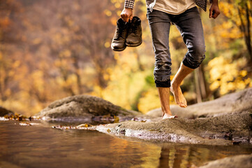 Male hiker crossing the stream barefooted walking by the creek with his shoes in hand adventure...