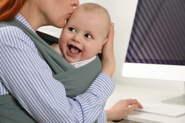 Mother holding her child in sling (baby carrier) while using computer at workplace