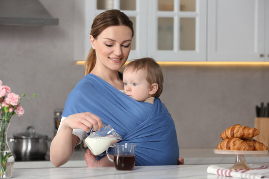 Mother adding milk into cup of drink while holding her child in sling (baby carrier) in kitchen